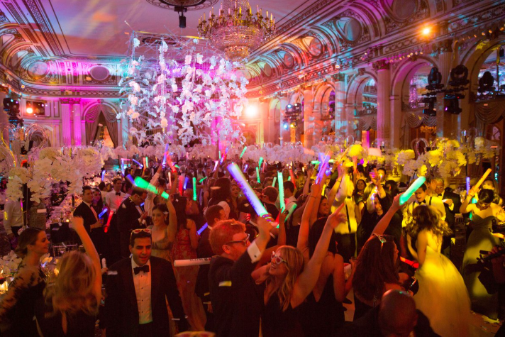 A bride and groom exchange vows under a floral arch on a beach, with the ocean and a boat in the background.