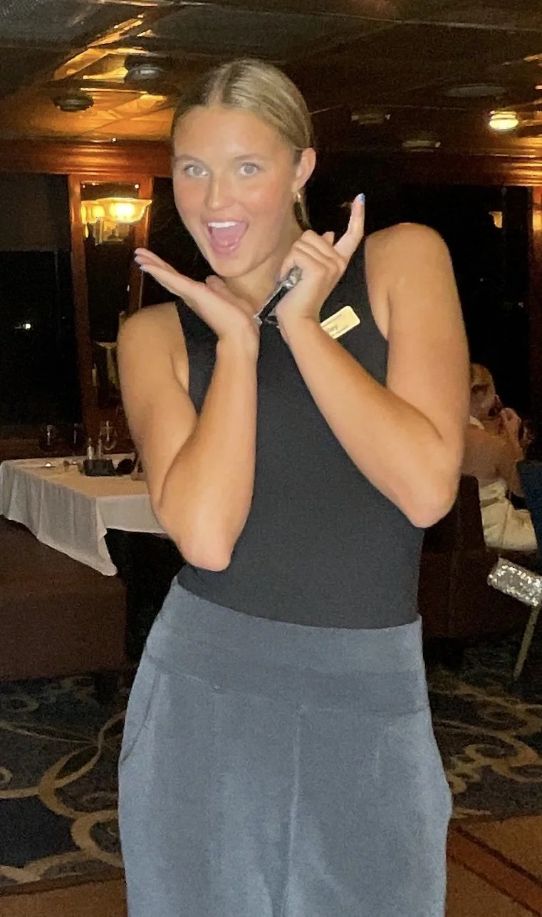 A woman with blonde hair in a bun, wearing a black sleeveless top and a gray skirt, smiling and posing with her hands framing her face inside a dimly lit restaurant or event space.