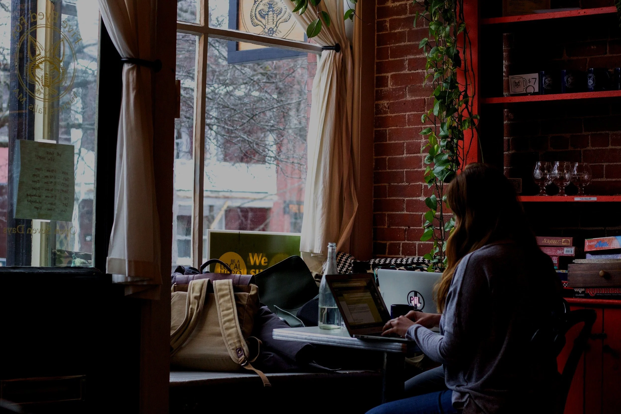 Woman working on a laptop in a cozy cafe with brick walls, plants, and large windows.
