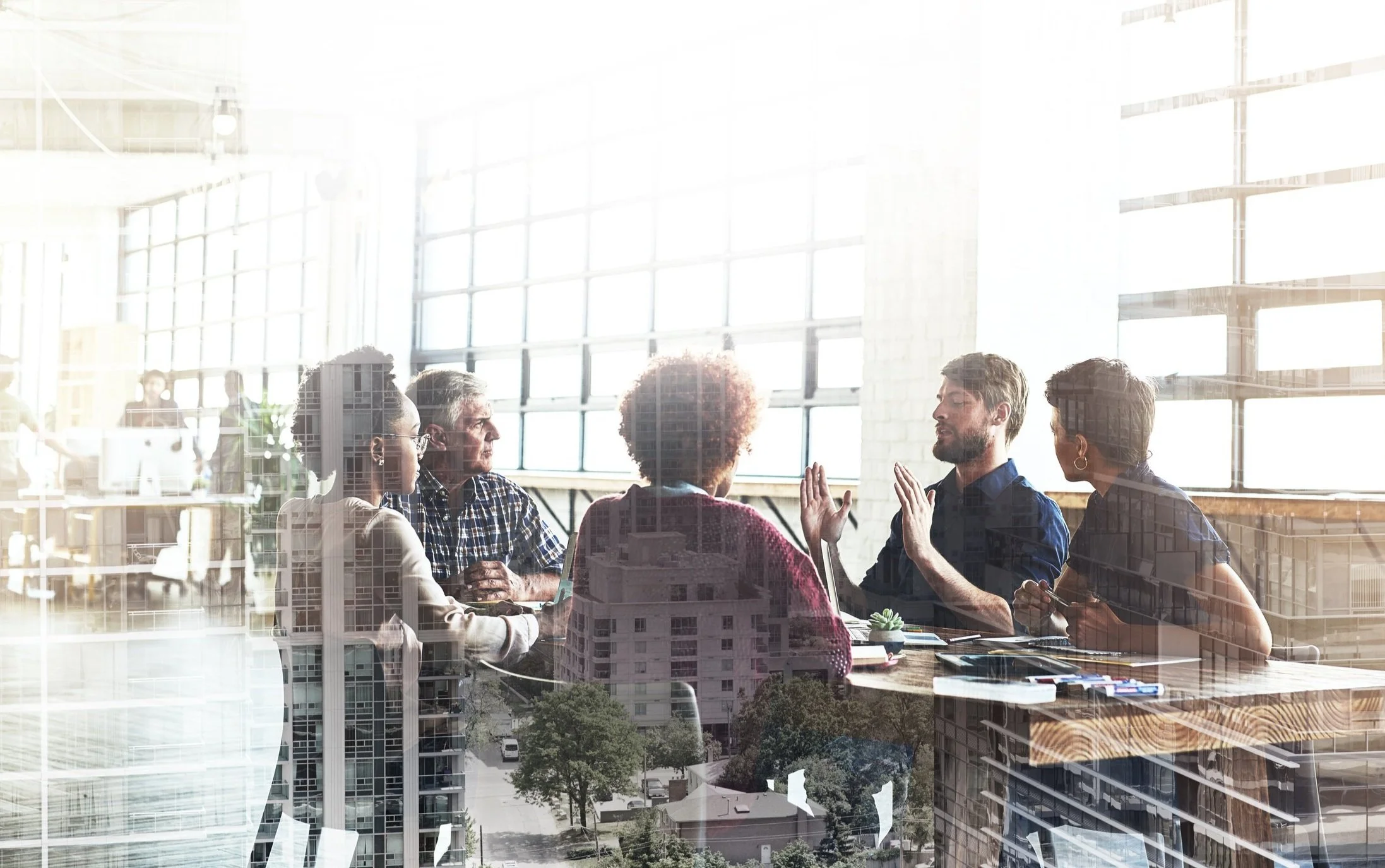 Double exposure of business meeting and urban landscape, featuring five people sitting around a table in a bright office setting with large windows, overlaid with city buildings.