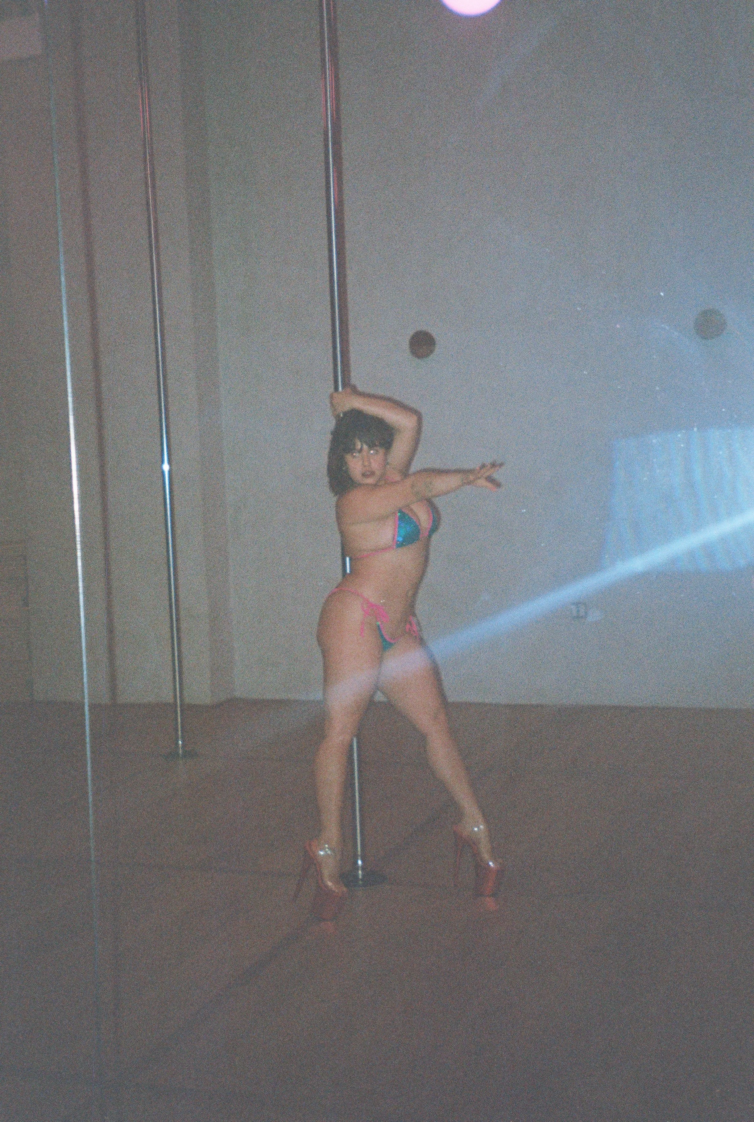 A woman in high heels and colorful lingerie, dancing and posing with a pole in a room with plain walls and a reflective floor.