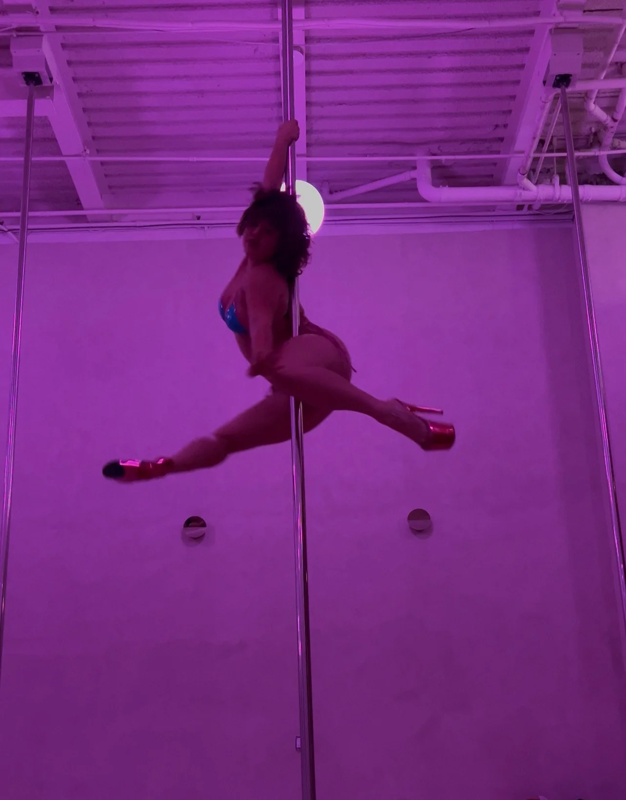 A woman performs a pole dance split maneuver on a pole, with purple lighting in the background.