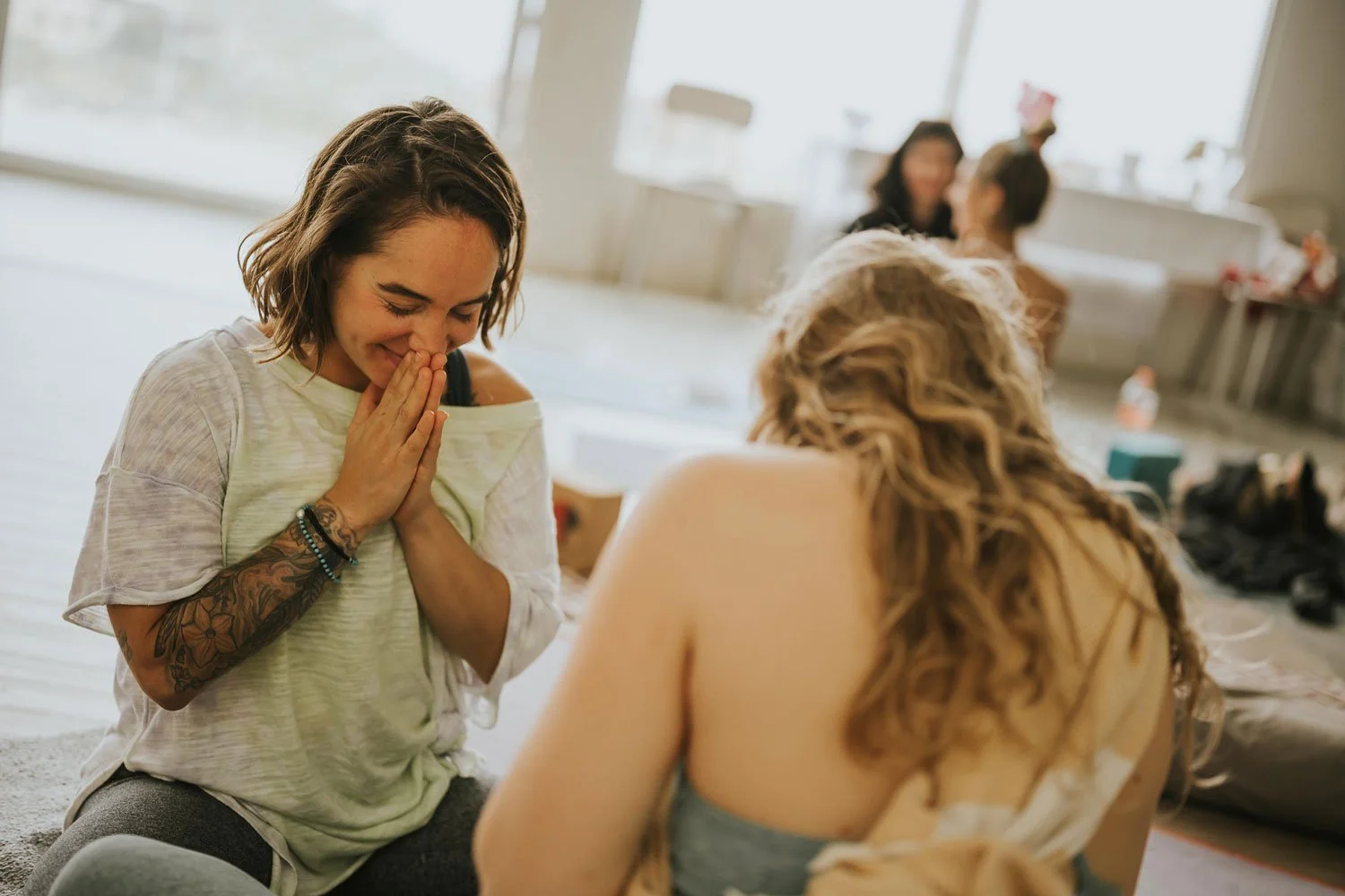A woman with tattoos on her arm and a joyful expression holds her hands to her face while kneeling, speaking with another woman with curly hair at a casual indoor gathering.