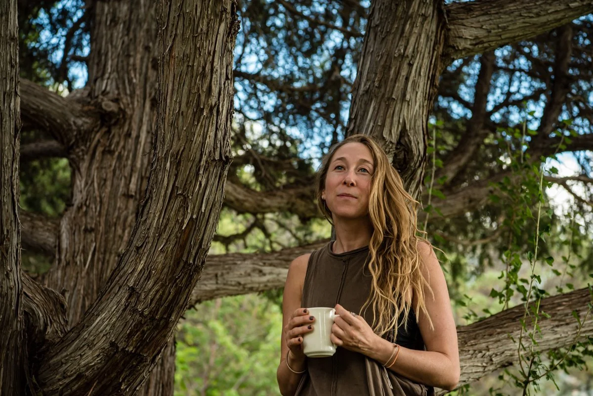 A woman with long blond hair stands outdoors holding a white mug near a large tree with textured bark and thick branches, surrounded by greenery.