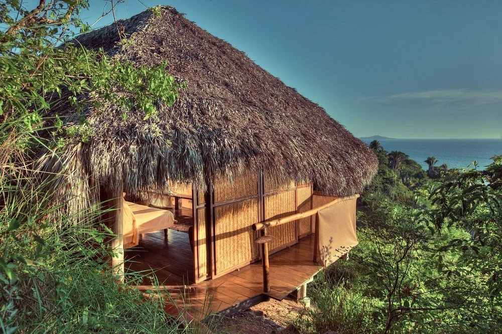 A thatched open-sided cabana raised on stilts at the edge of the jungle, its wooden deck looking out over treetops to the blue Pacific
