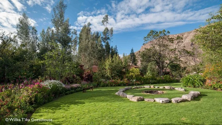 A lush green garden with a fire pit surrounded by large stones, colorful flowers, and trees under a partly cloudy sky, with mountains in the background.