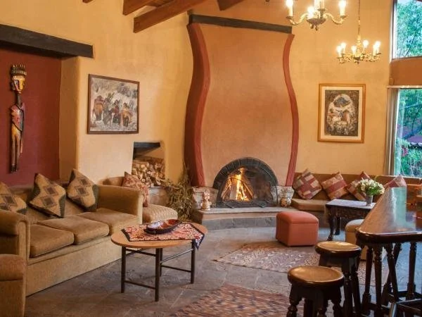 Living room with a large fireplace, beige sofa with patterned pillows, coffee table, artwork on walls, chandelier, and window showing greenery outside.