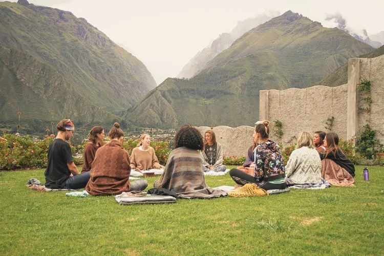 A group of people sitting on the grass in a circle, engaging in a discussion or meditation session, with mountains in the background.