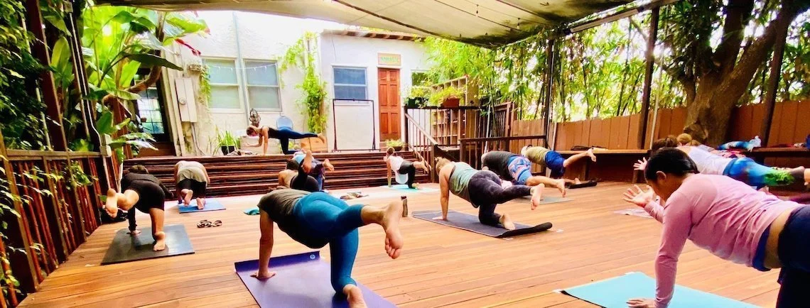 Group of people doing yoga on outdoor wooden deck under a canopy, surrounded by greenery.