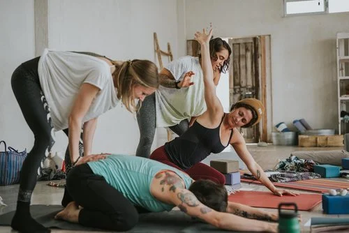 Two women practicing yoga on mats with a woman assisting them in a bright, industrial-style studio.