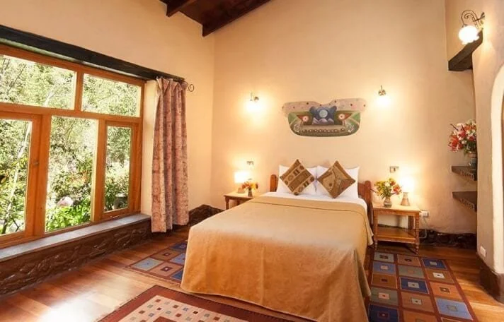 A cozy bedroom with a large window, beige walls, and a wooden ceiling beam. It has a double bed with beige bedding, two pillows, and a colorful decorative pillow. On each side of the bed, there are small bedside tables with lamps and flower arrangements. Above the bed, there is a decorative wall hanging. The room has a patterned area rug and a shelf with flowers on the right side.