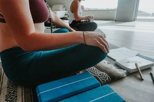 A person practicing yoga or meditation seated cross-legged on a mat in a sunny room, with a notebook and pen nearby, and another person sitting in the background.