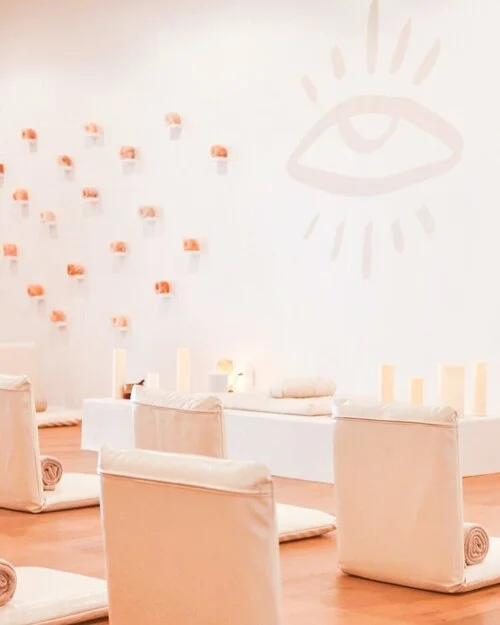 A minimalistic room with a white wall, tan chairs, and floral decorations on the wall, featuring a large eye symbol drawn in the upper right corner.
