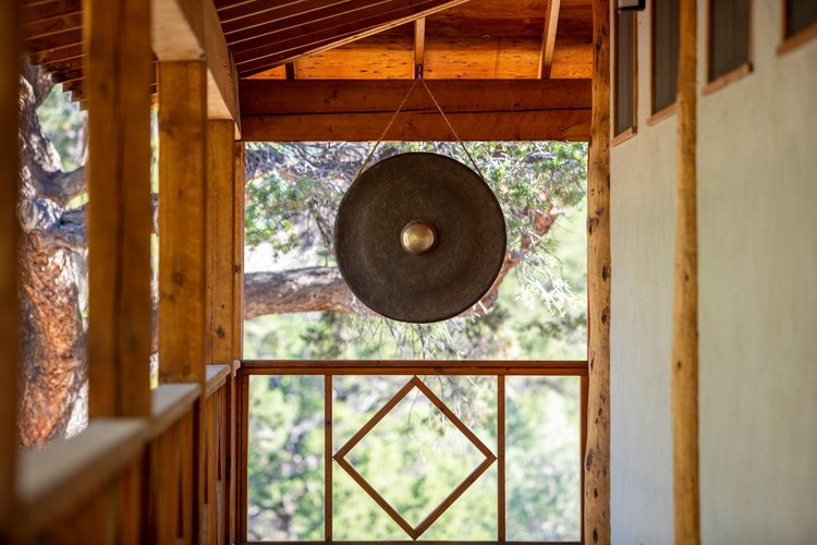 A balcony with wooden railing and ceiling, featuring a large metal gong hanging from the ceiling, with trees visible in the background.