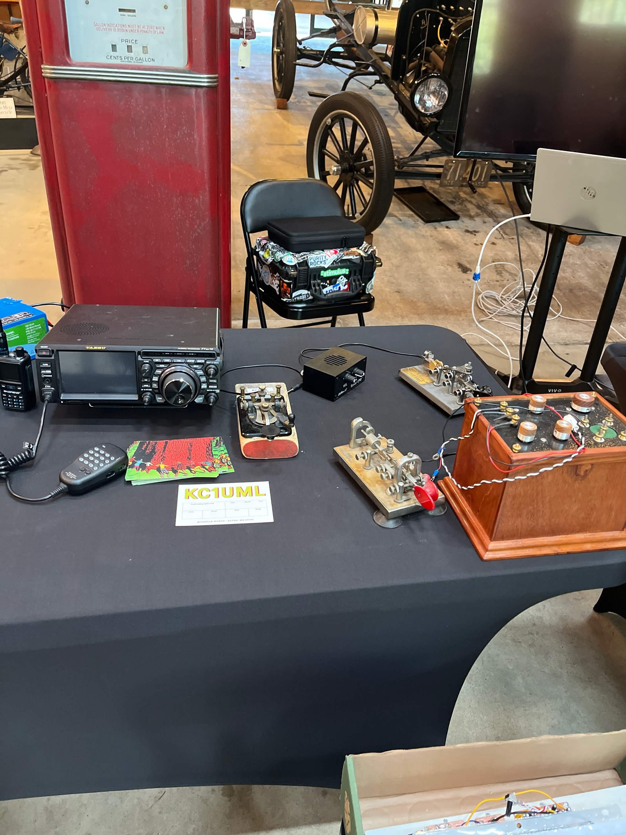 A table with electronic equipment, including radios, gadgets, and a small wooden box, is set up in a room with a vintage red gas pump, a black folding chair with a box of supplies, and an antique vehicle with a large wheel in the background.