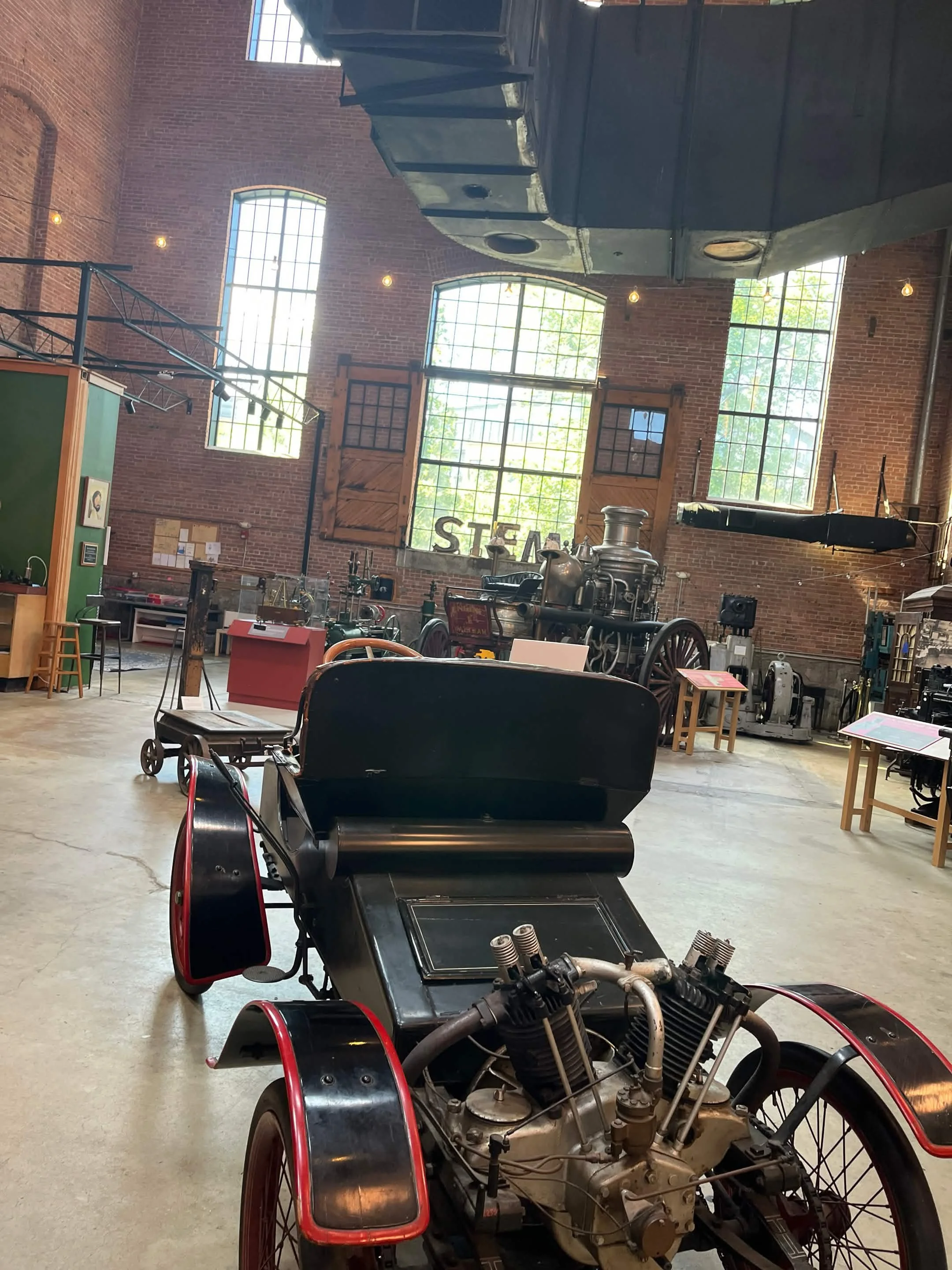 An indoor exhibit space with vintage machinery, large brick walls, tall windows, and a wooden sign reading 'STEAM'.