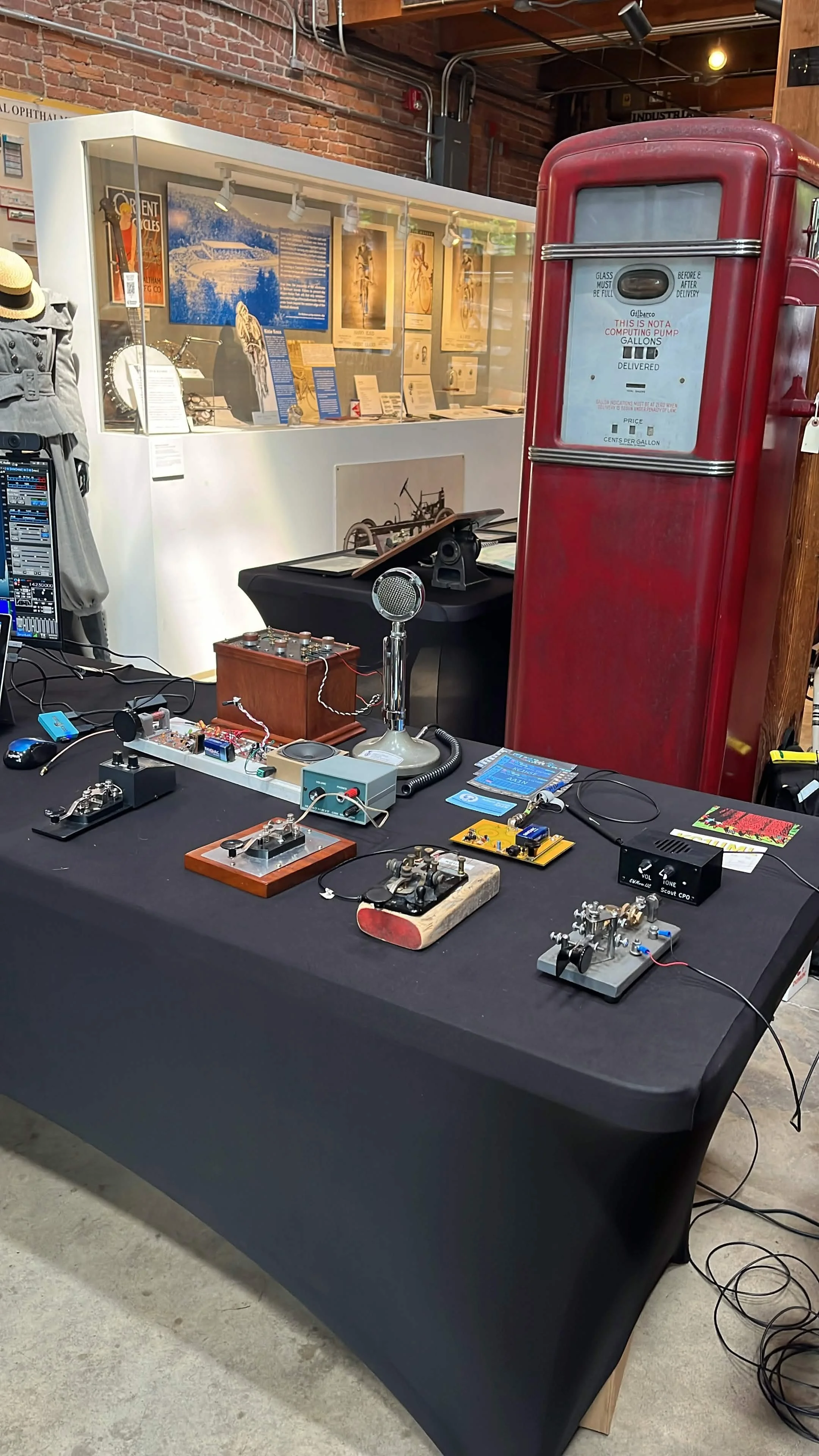 A display of vintage electronic devices and scientific experiments on a black table, including small metal or electronic components, a vintage microphone, and circuit boards, with a red vintage gas pump and framed informational displays in the backgr