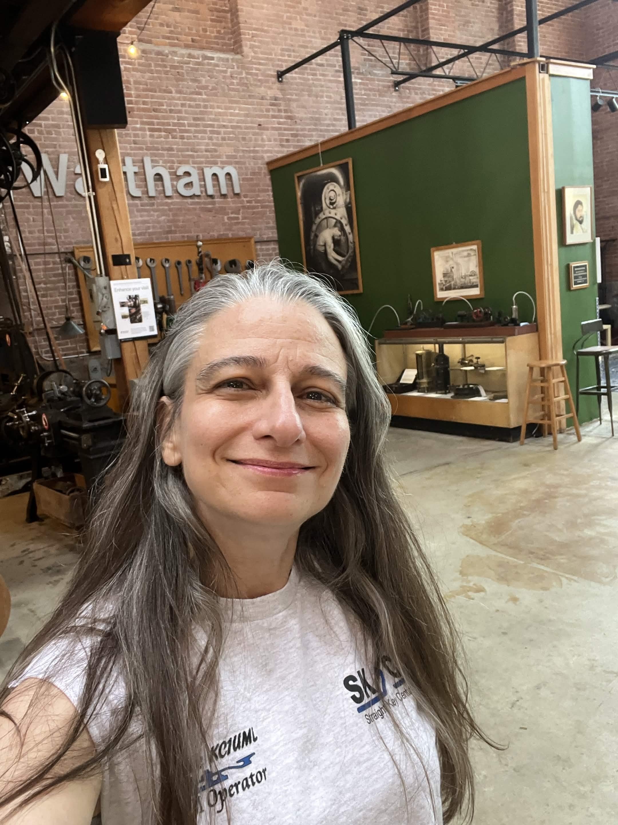 A woman with long gray hair smiling in an industrial-style space with exposed brick walls, artwork on the green partition, and various machinery or tools in the background.