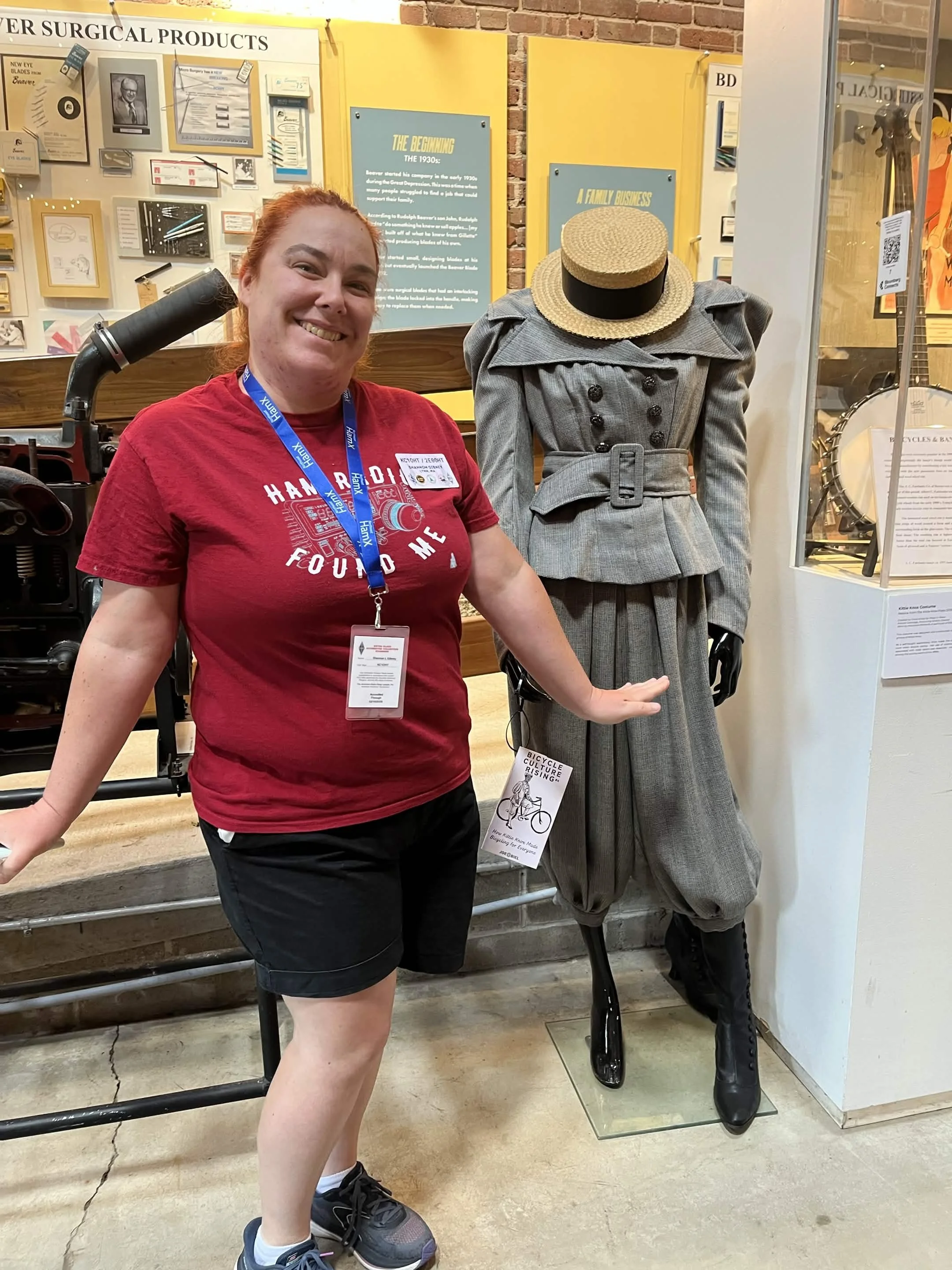 A woman with red hair smiling and wearing a red t-shirt and black shorts, standing inside a museum next to a mannequin dressed in vintage gray women's suit with a straw boater hat, in front of yellow informational displays about surgical products and
