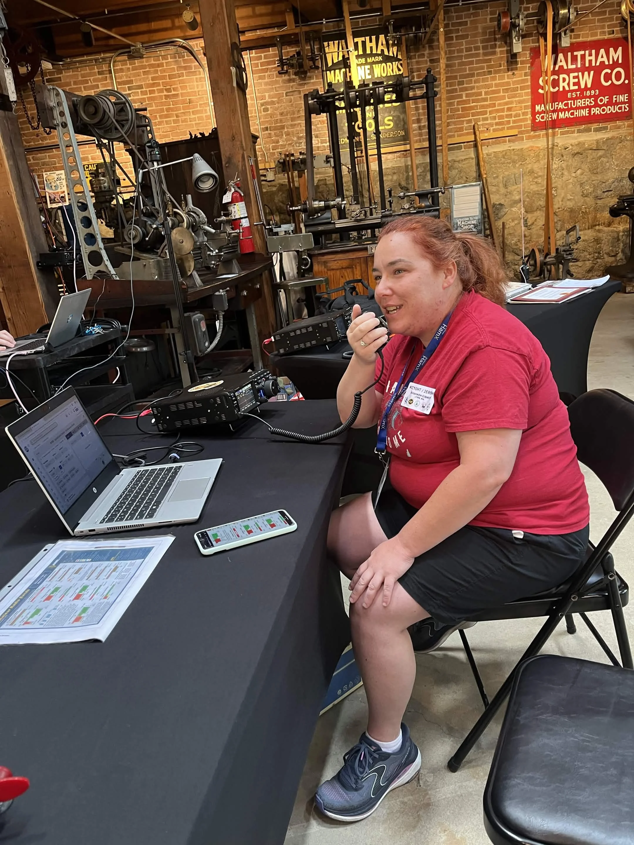 A woman with red hair, wearing a red t-shirt, black shorts, and gray running shoes, is sitting at a table with radio equipment, listening to a walkie-talkie. Behind her is a brick wall with machinery and vintage signs related to machine works and scr