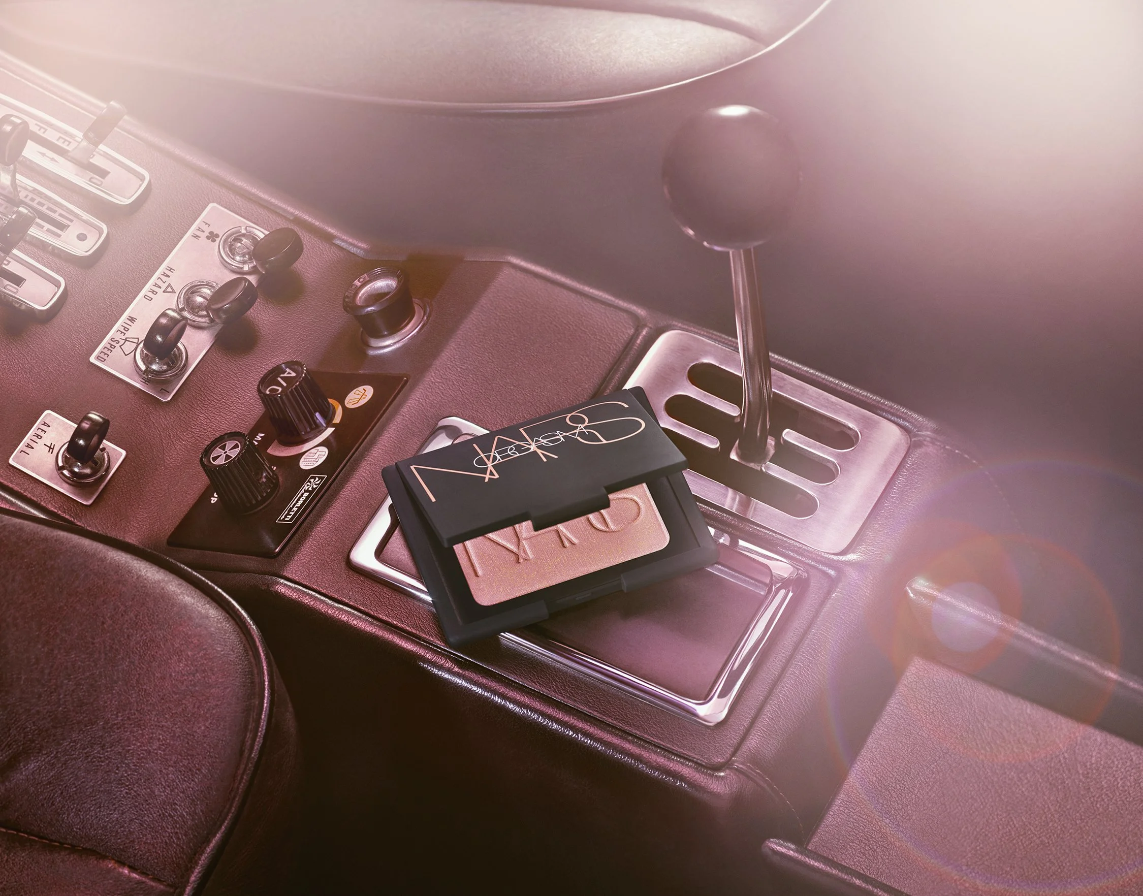 A NARS compact blush and a car's manual gear shift lever in a vintage vehicle interior with control knobs and switches on the dashboard.