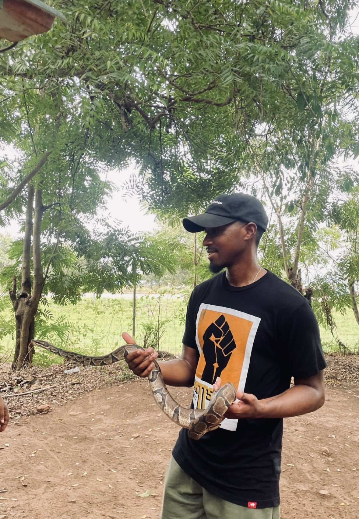 A man wearing a black cap and t-shirt holding a large snake outdoors, with green trees and a wire fence in the background.