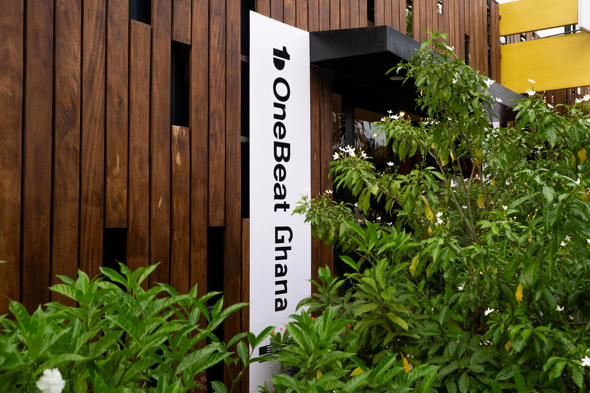 A white vertical sign with black text reading '1st one B Eat G Nana' is mounted on a wooden wall surrounded by green plants and white flowers.