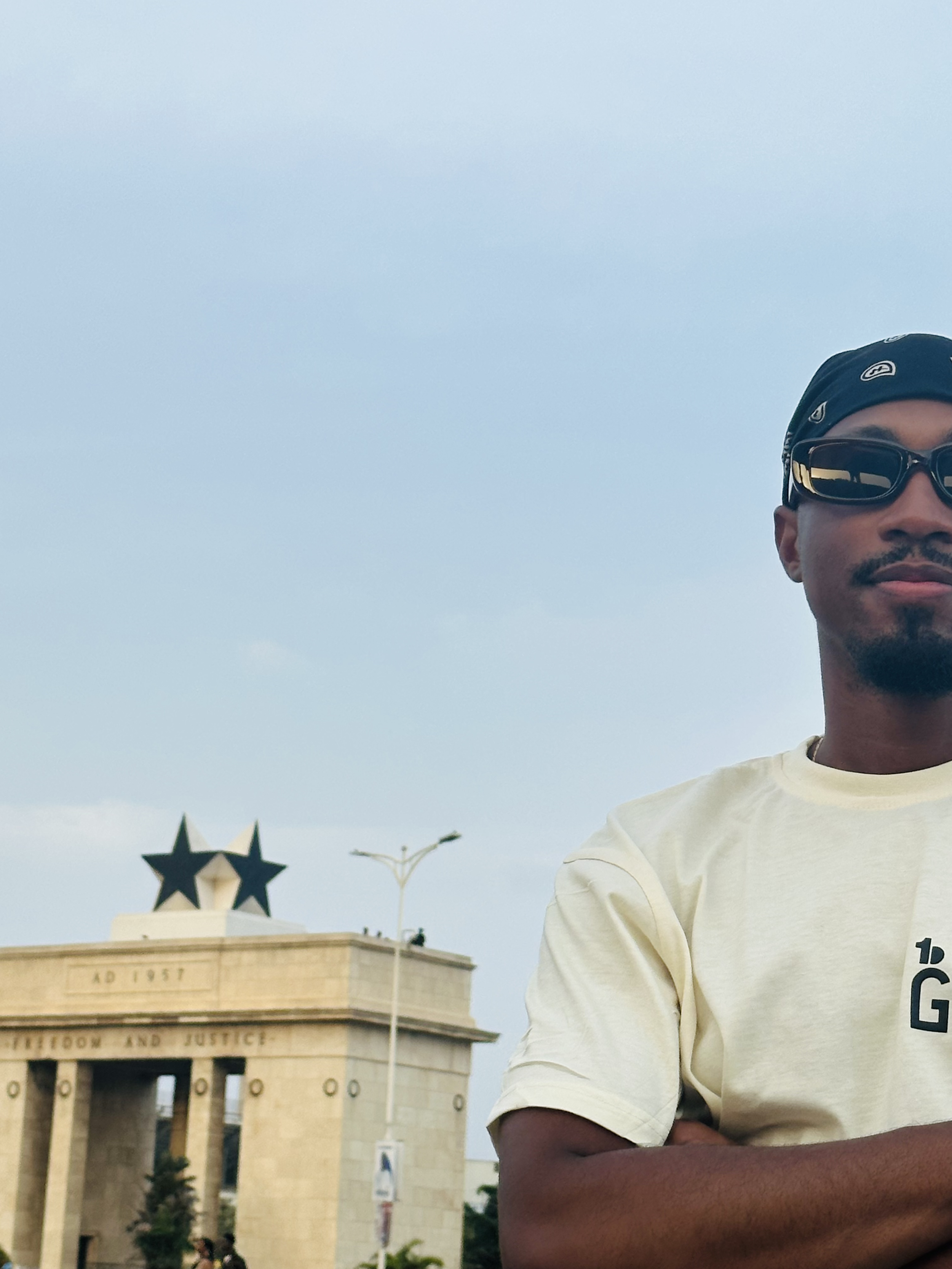 A man wearing sunglasses and a bandana in front of the Victory Gate at Red Square with a star on top, blue sky, and some streetlights.