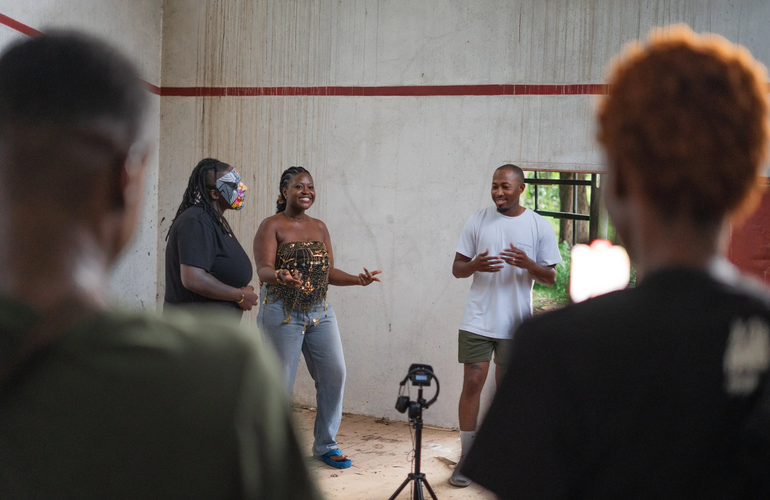 Three people speaking to an audience indoors, with two women and one man standing and speaking, while the audience watches. The background shows a concrete wall and a window.