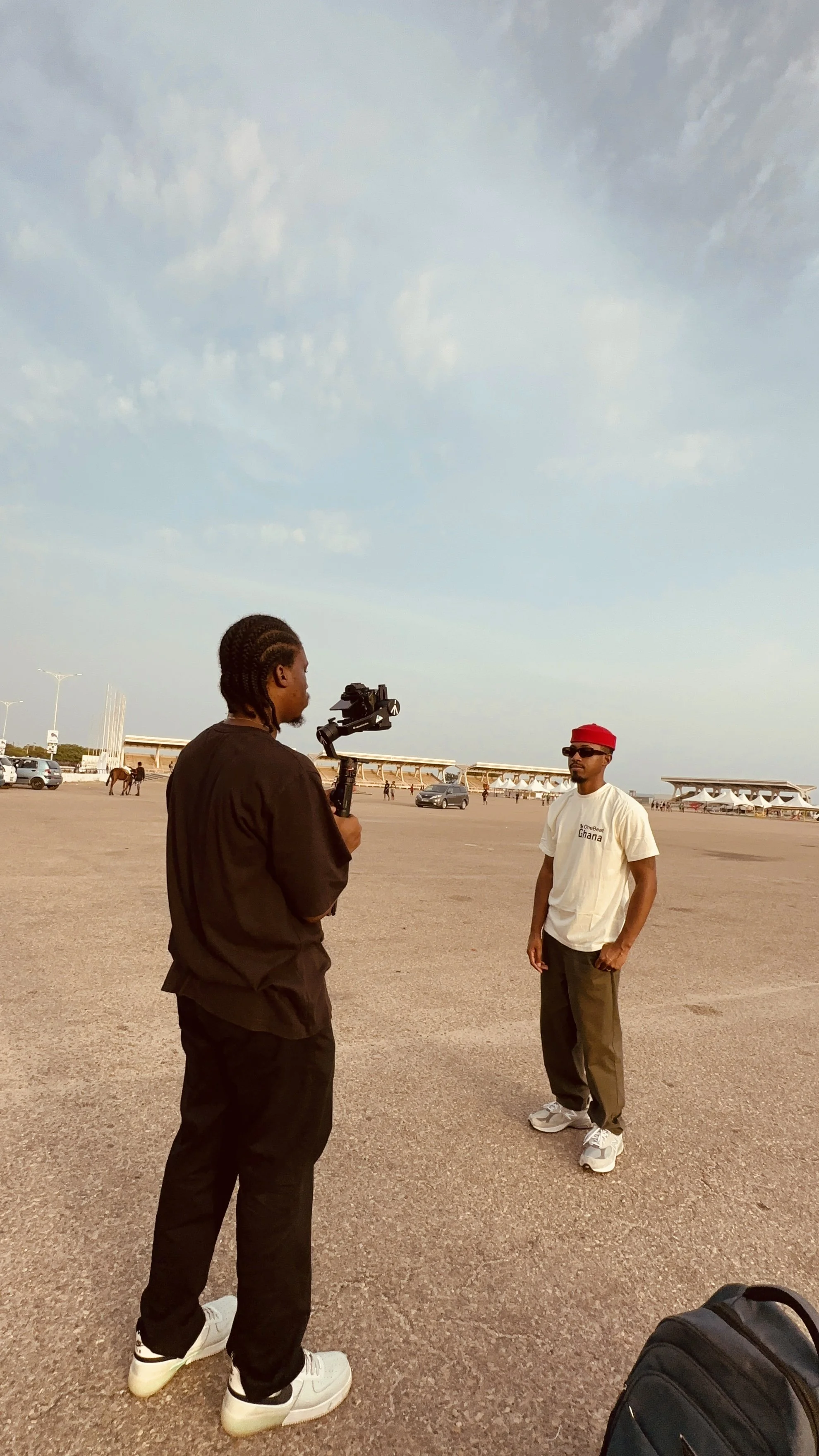 A person holding a camera filming another person wearing sunglasses, a white T-shirt, and brown pants in an open outdoor space with parked cars and horses in the background.