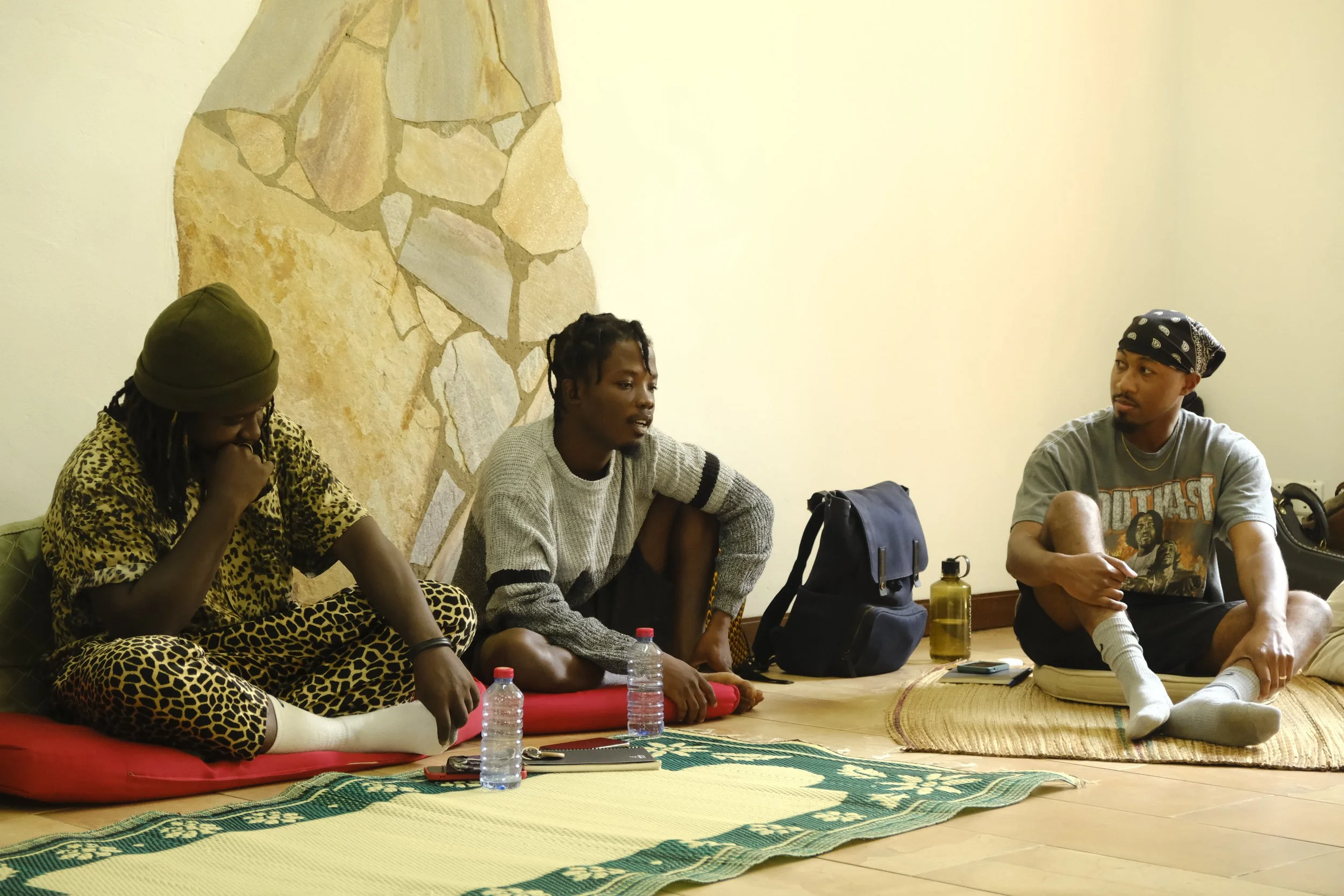 Three young men sitting on the floor in a room with a decorative stone wall. They are engaged in a conversation. The man on the left is wearing a leopard-print outfit and a green beanie, the man in the middle is wearing a gray sweater and black short
