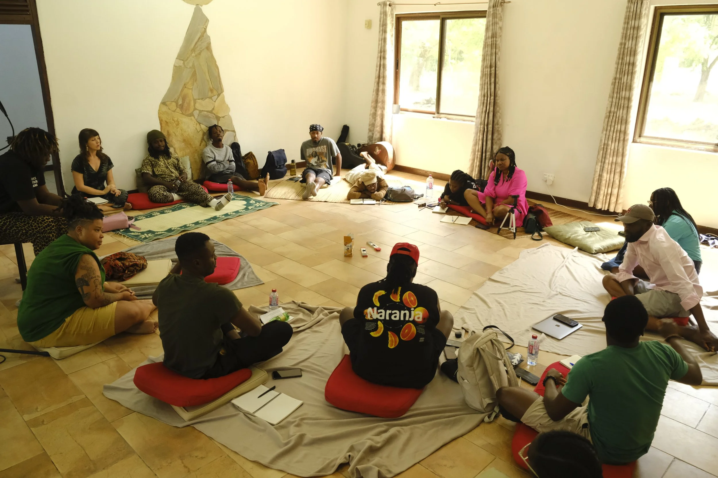 A group of people seated on mats and cushions in a circle in a bright room with large windows.