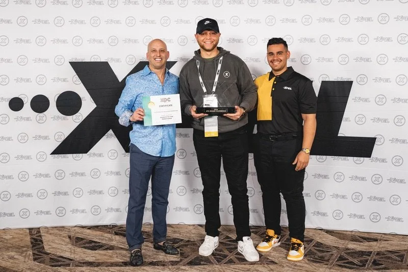 Three men standing in front of a branded backdrop, with one holding a certificate and another holding a trophy at an event.