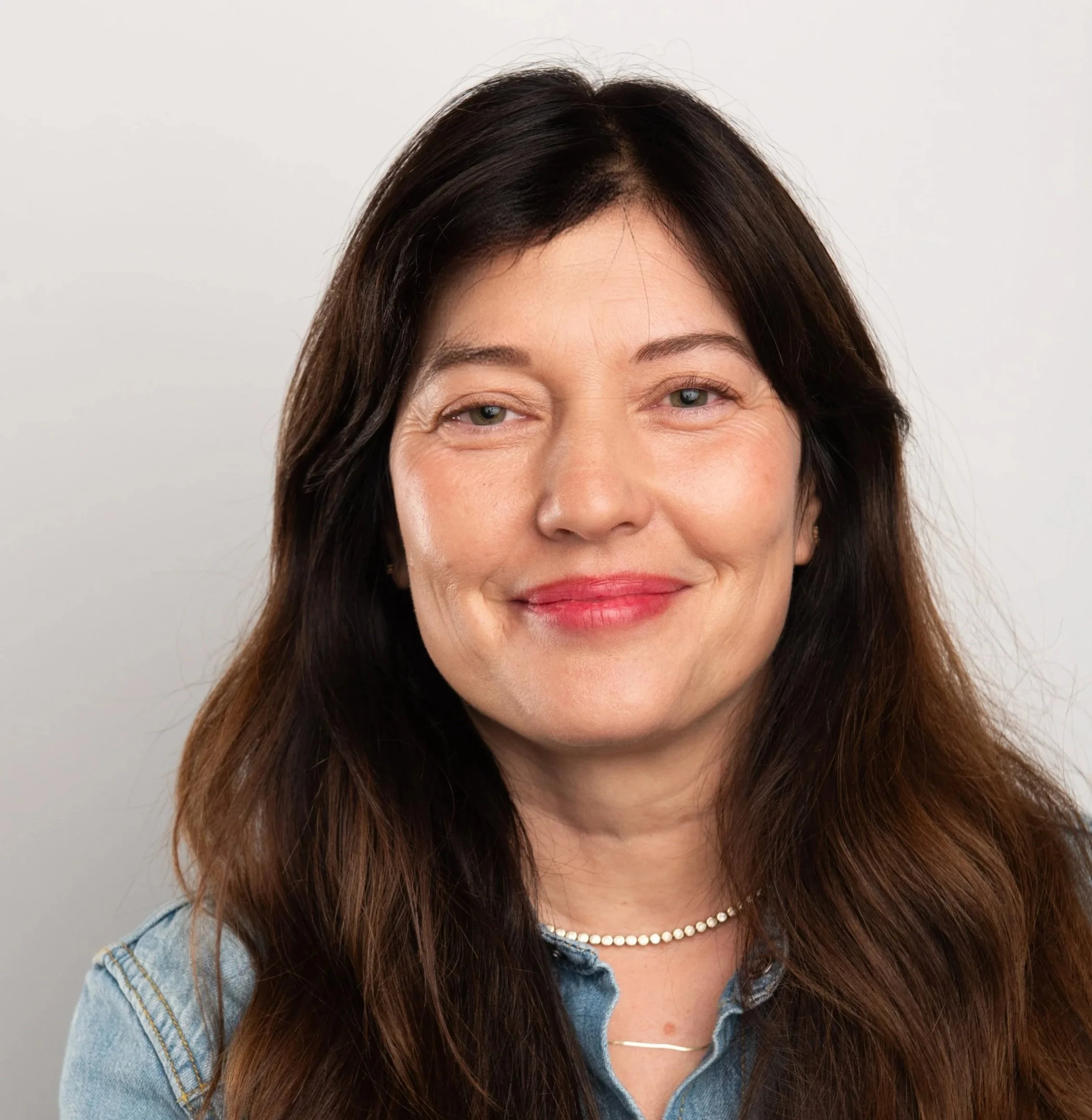 Close-up portrait of Dr. Regina Vanburg with dark brown hair, light skin, wearing a pearl necklace and denim jacket, smiling against a plain white background.