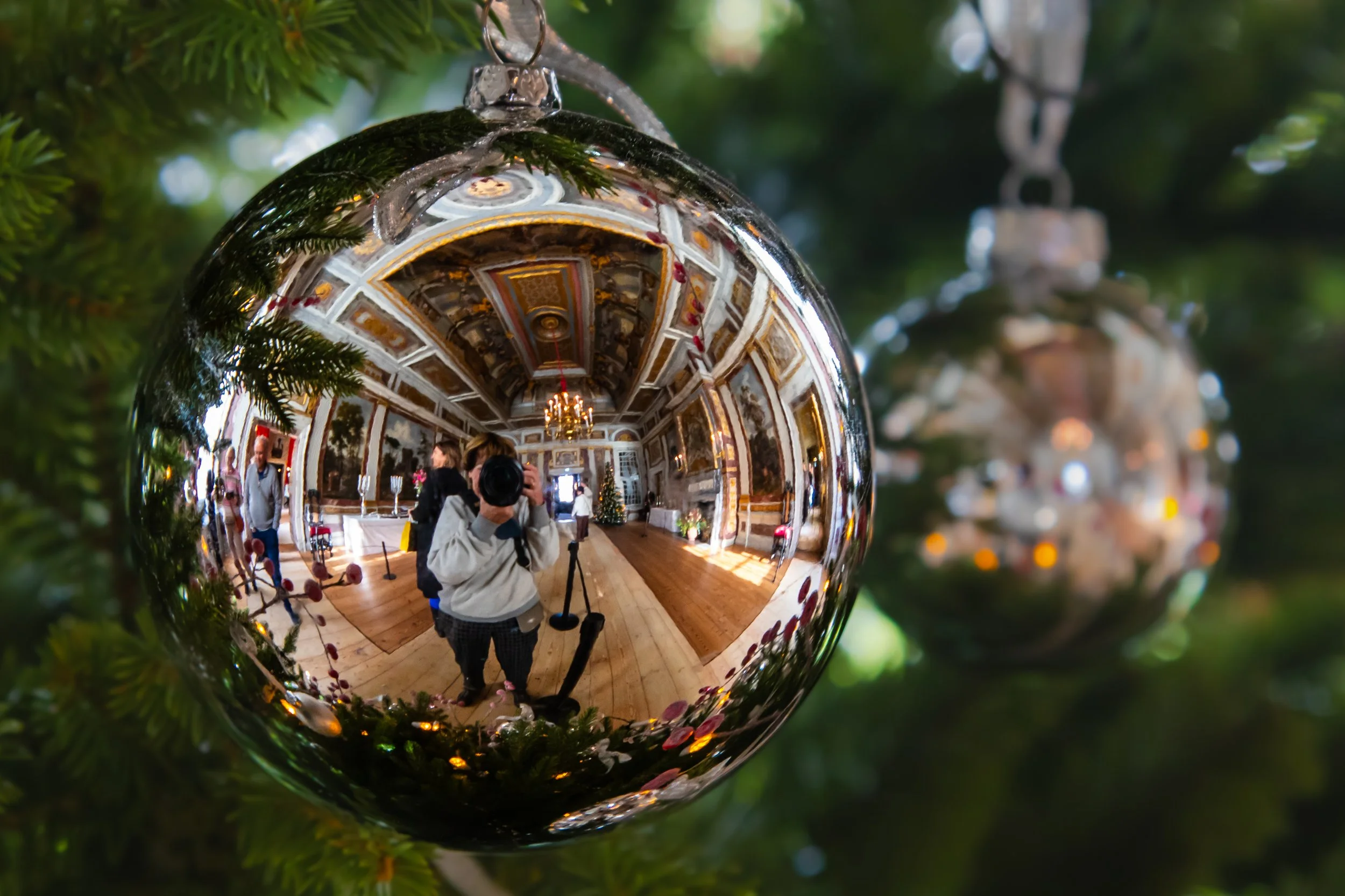 Reflectie in een kerstbal van een fotograaf die een kamer met ornamenten en mensen fotografeert, omgeven door een boom met andere kerstballen.