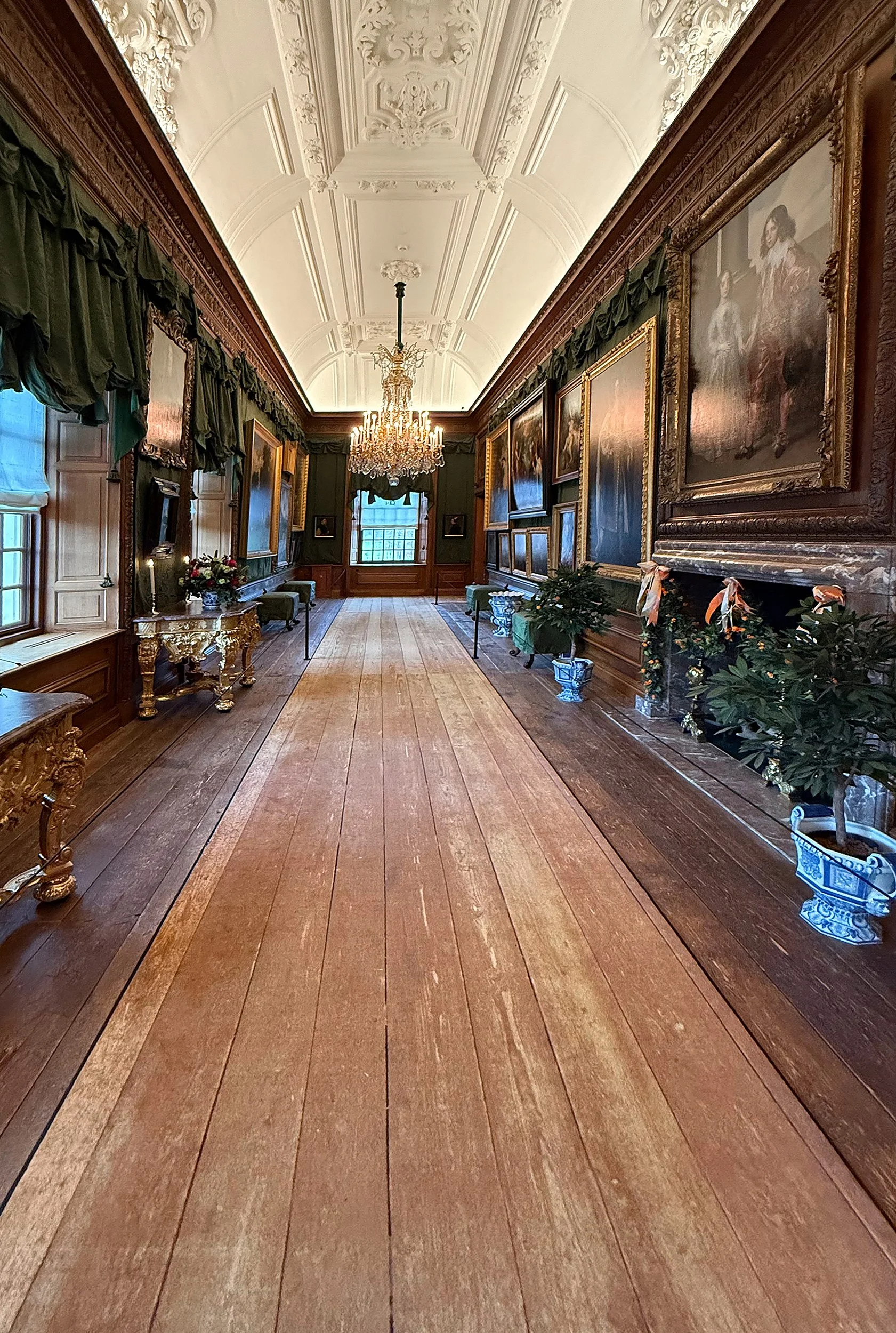 Elegante zaal met houten vloer, laden wanden, grote schilderijen met gouden lijsten, groene gordijnen, een kristallen kroonluchter en planten in blauwe en witte potten.