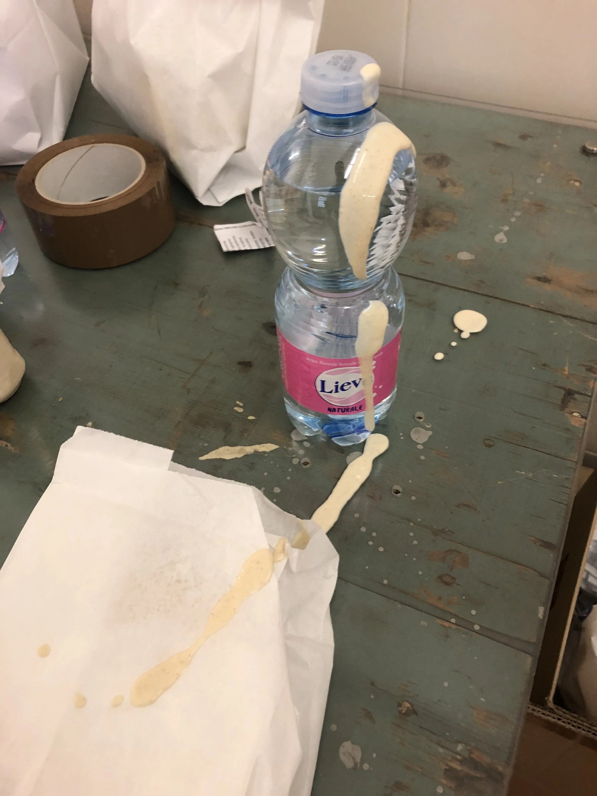 Una bottiglia d'acqua con etichetta rosa e del liquido beige fuoriuscito, su un tavolo verde, accanto a un rotolo di nastro adesivo e una busta di carta bianca.