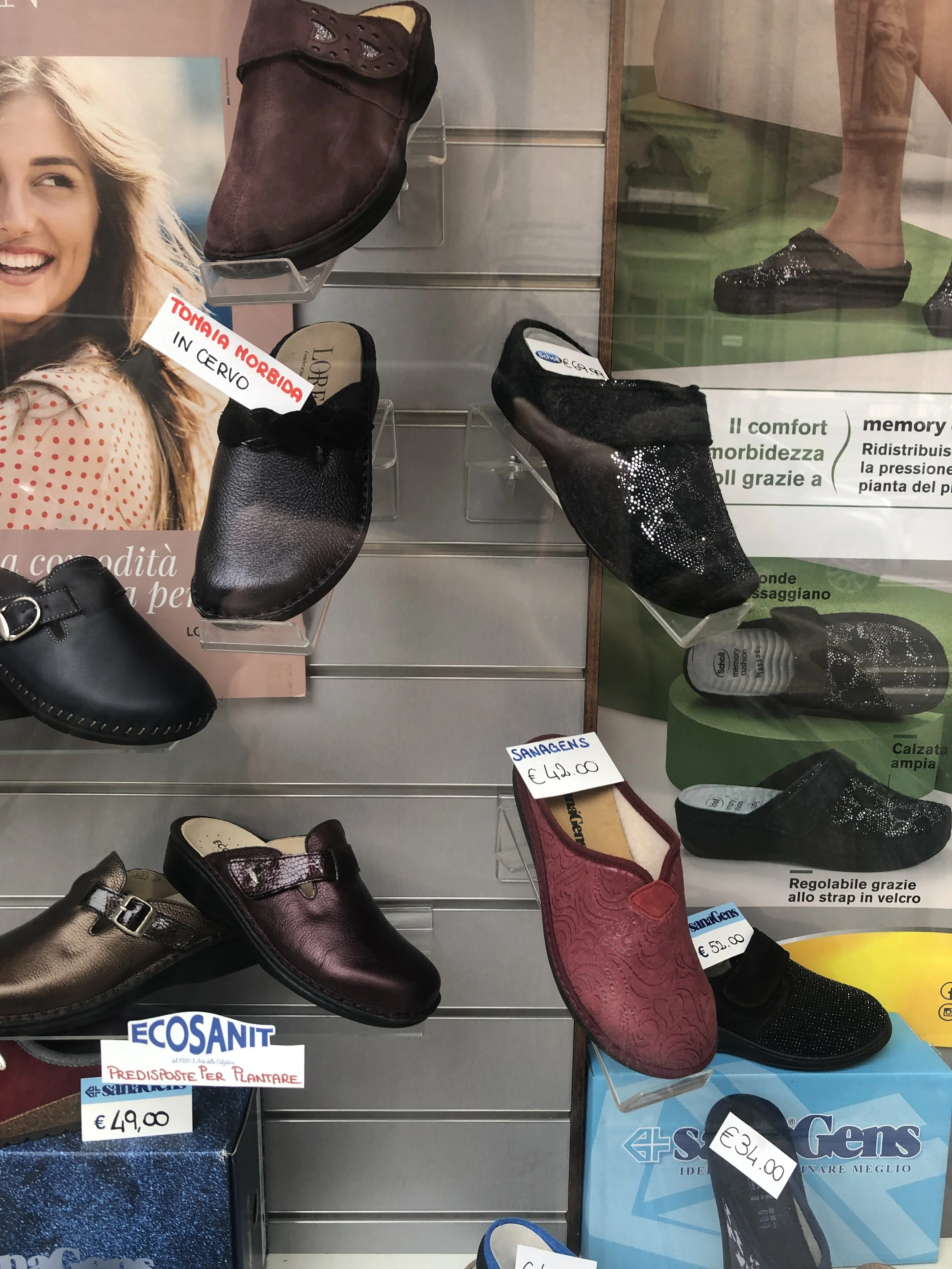 Vetrina con vari modelli di calzature ortopediche, alcune con etichette che mostrano i prezzi, come "€49,00" e "€34,00". Include marchi come Ecosanit e Sanagens. Ci sono anche cartelli pubblicitari per il comfort e la morbidezza.