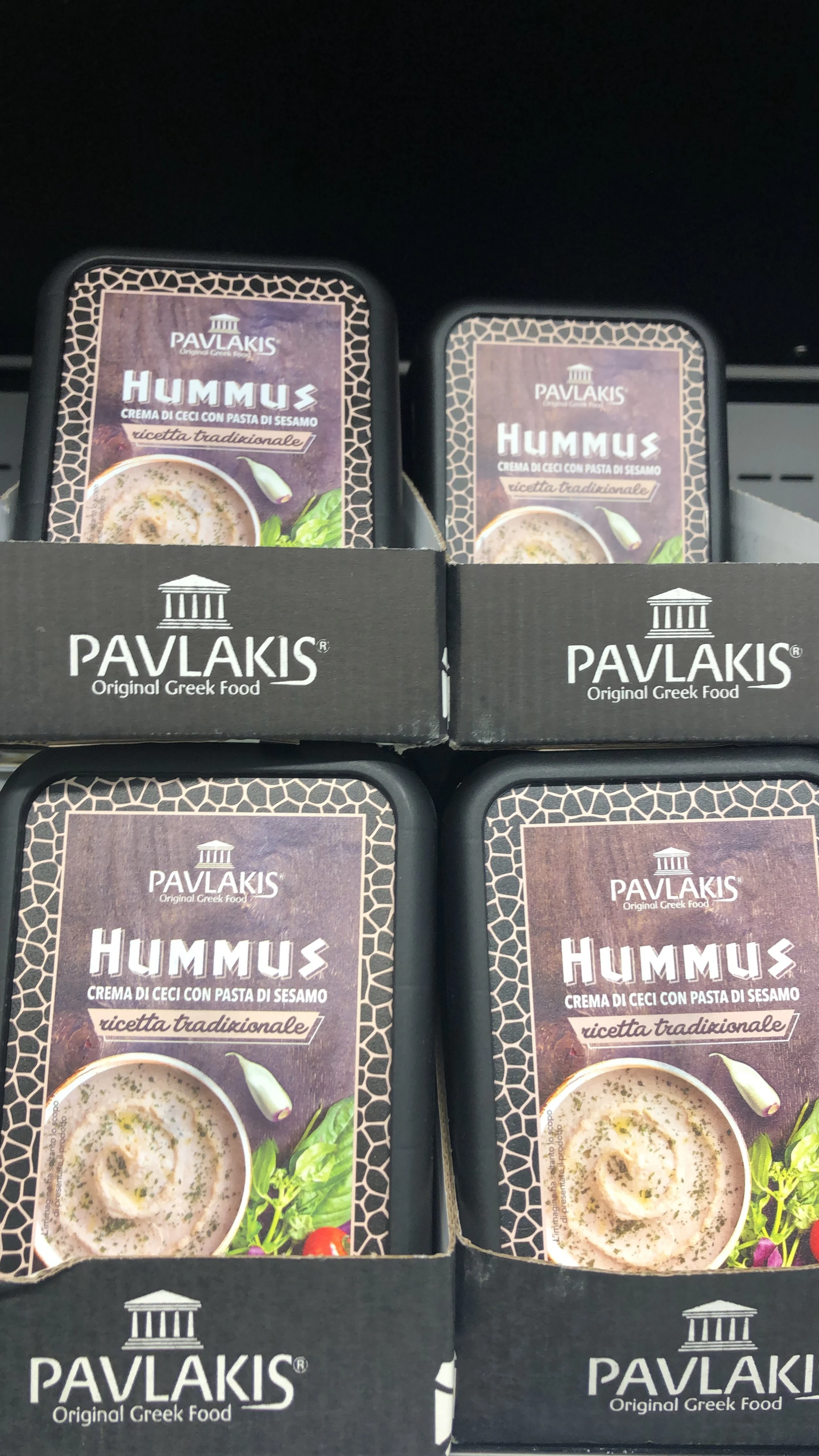 Contenitori di hummus Pavlakis in un supermercato, etichettati "crema di ceci con pasta di sesamo" e "ricetta tradizionale". Immagini di hummus con verdure fresche sulla confezione. Prodotti posizionati su scaffali.