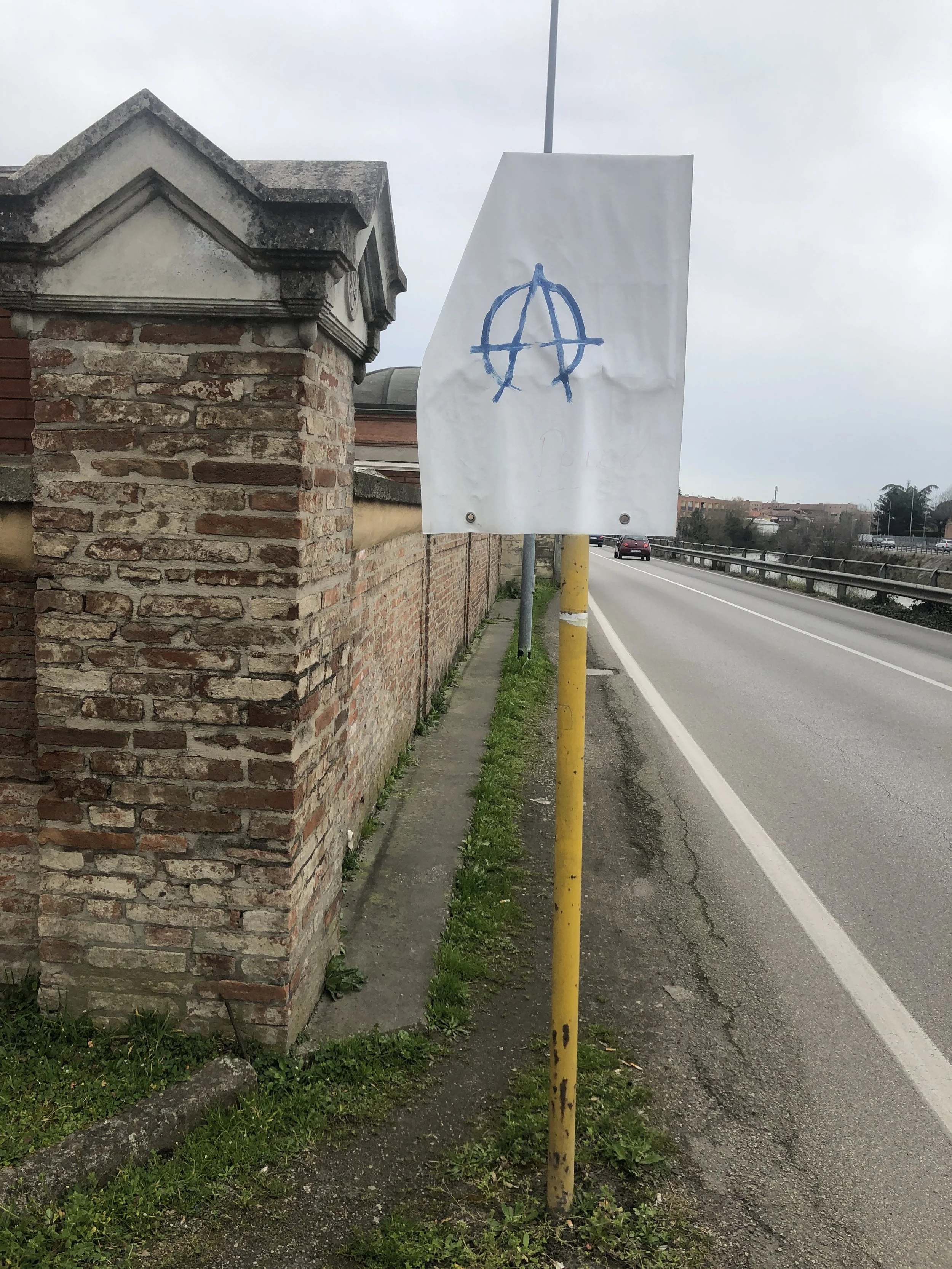 Cartello bianco con simbolo anarchico blu appeso a un palo giallo vicino a un muretto di mattoni su una strada.