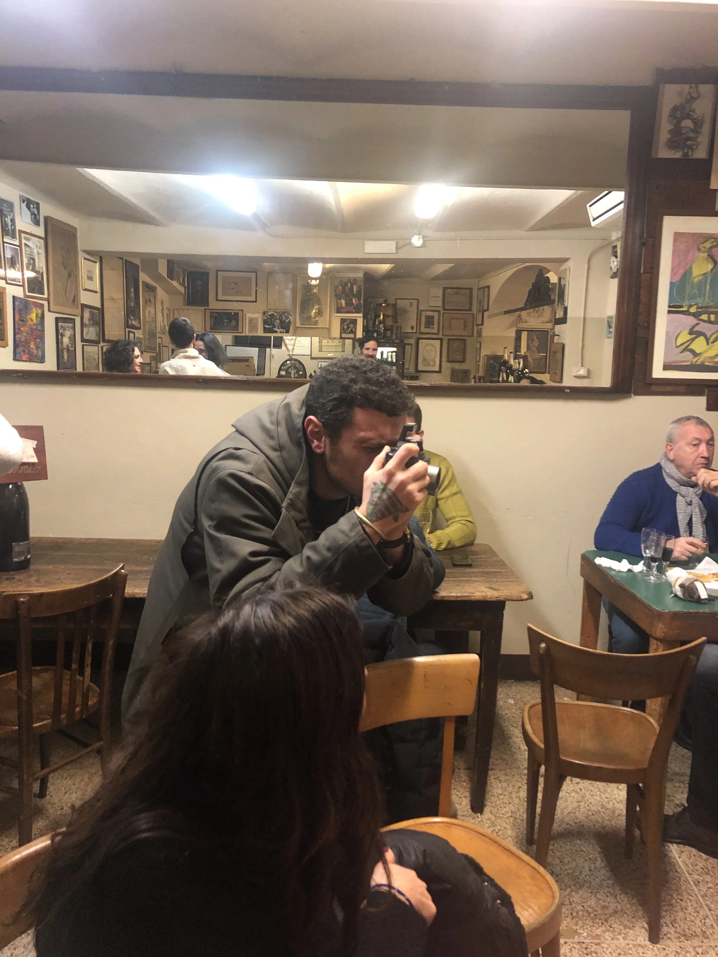 Una persona sta scattando una foto in un ristorante accogliente, con tavoli e sedie in legno. Ci sono diverse opere d'arte appese alle pareti e una grande specchiera riflette la scena. Altre persone sono sedute ai tavoli.