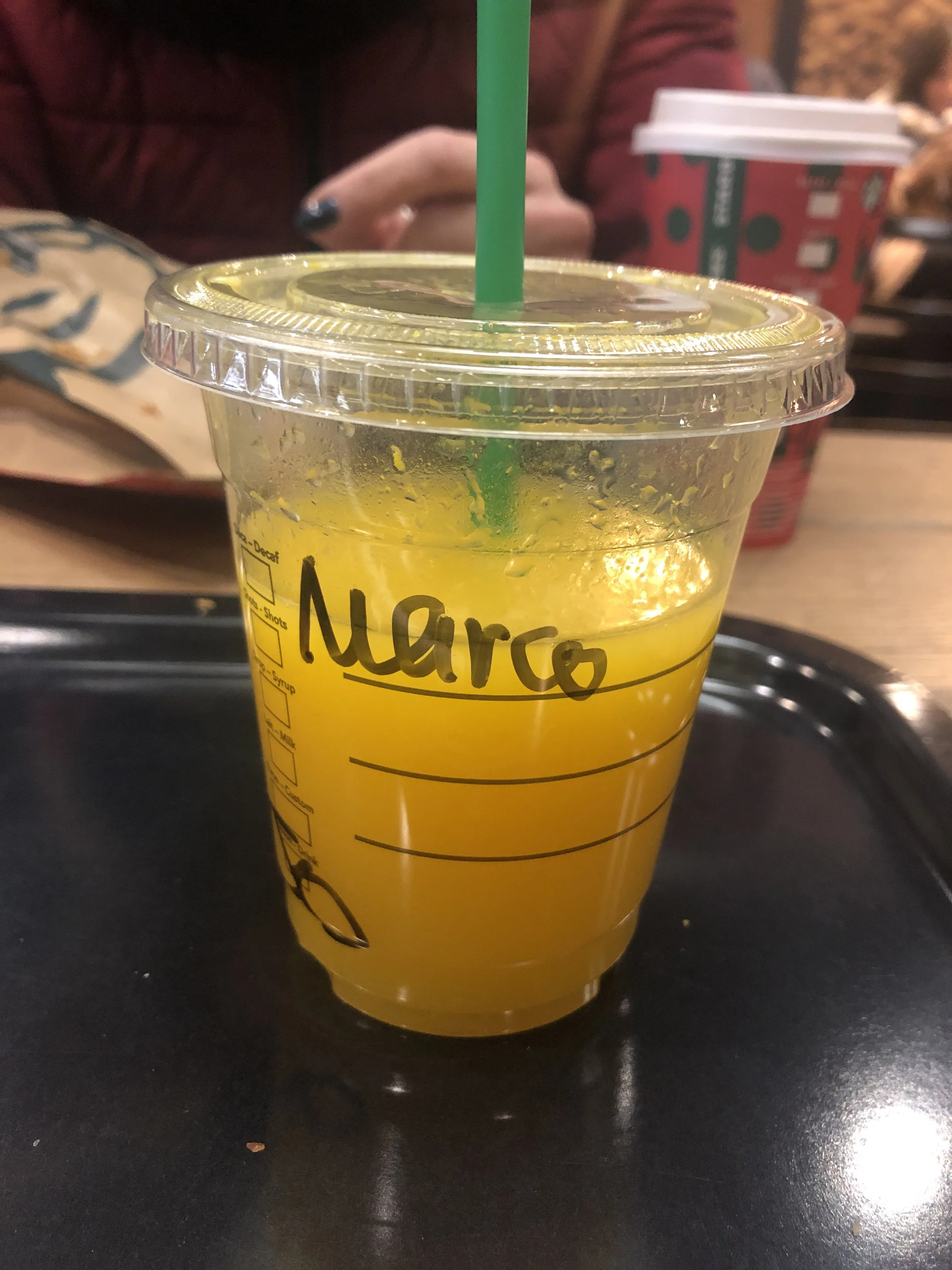 Bicchiere di plastica con succo d'arancia e cannuccia verde, con il nome 'Marco' scritto sul lato. Sfondo di una tazza e una persona.