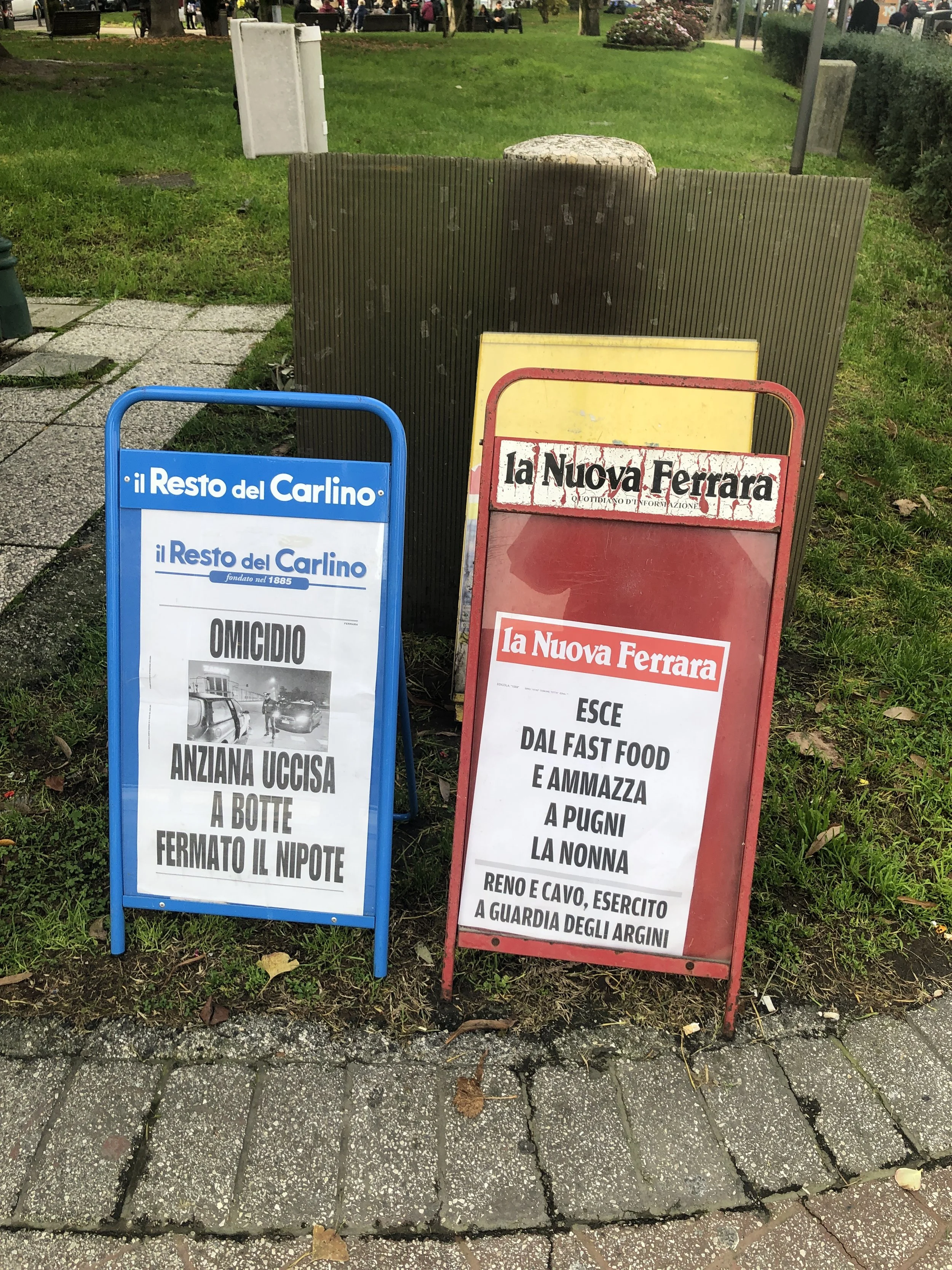 Due cartelli di giornali su un marciapiede. Il primo cartello è de "il Resto del Carlino" e riporta una notizia su un omicidio di un'anziana uccisa a botte, indicando che il nipote è stato fermato. Il secondo cartello è de "la Nuova Ferrara" e parla 