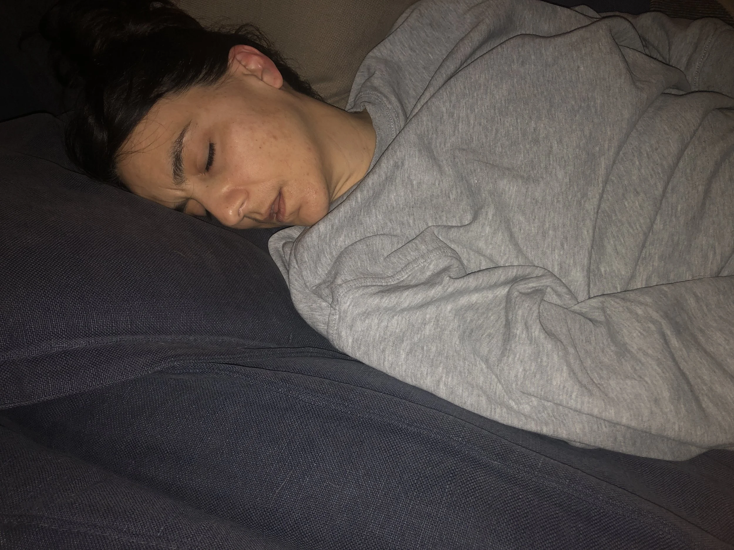 Persona addormentata su un divano, indossa una felpa grigia, appoggiata su un cuscino scuro.