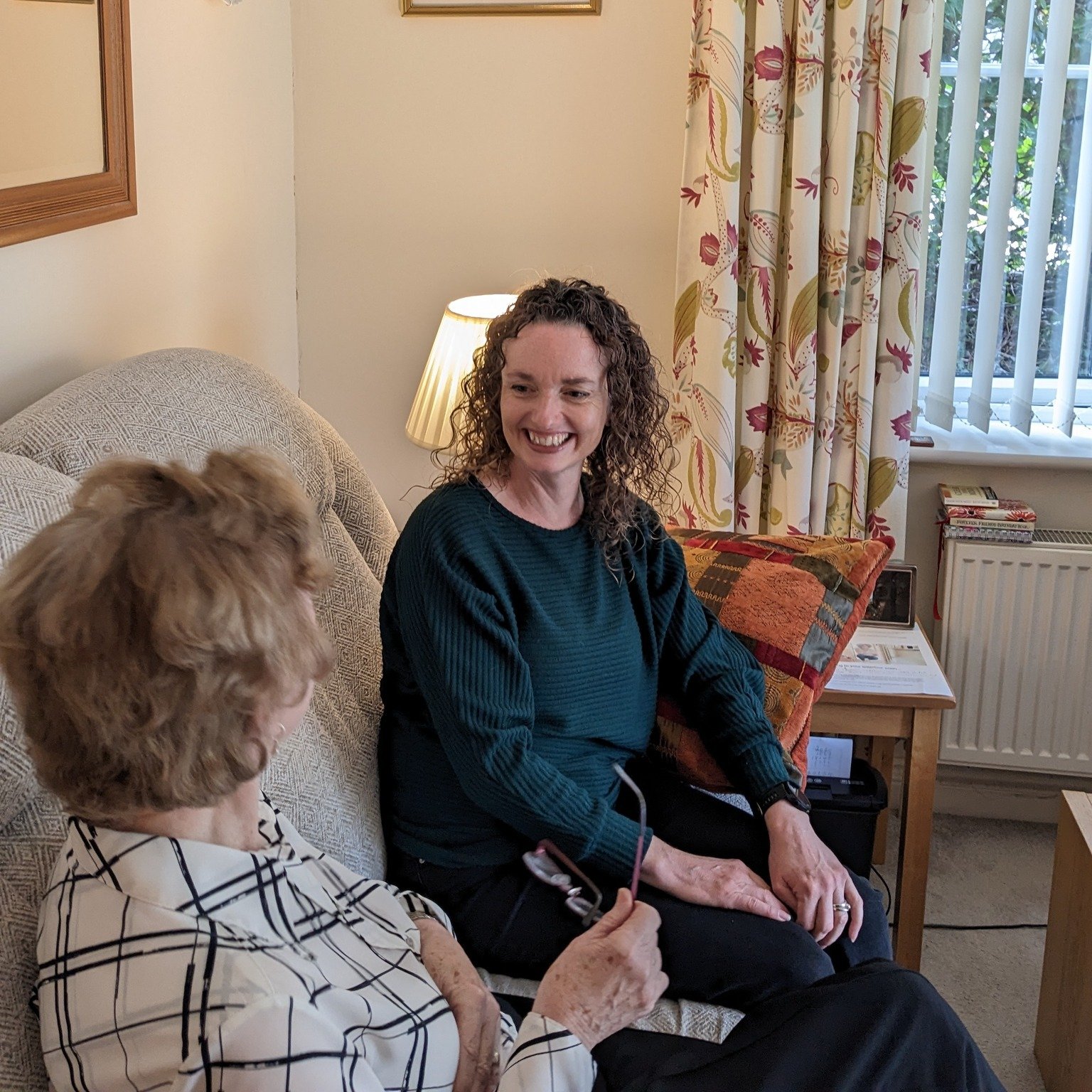 Two women sitting on a beige couch in a living room, engaging in conversation. One woman has curly hair and is smiling, while the other has short hair and holds glasses.