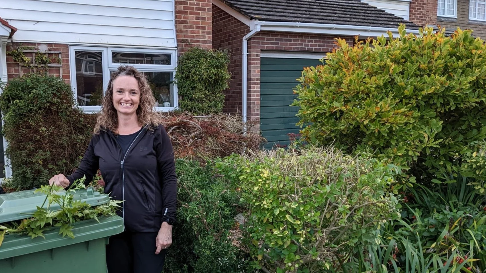 Smiling woman in black jacket standing in front of a garden with bushes and plants, house with brick walls and a window in the background.