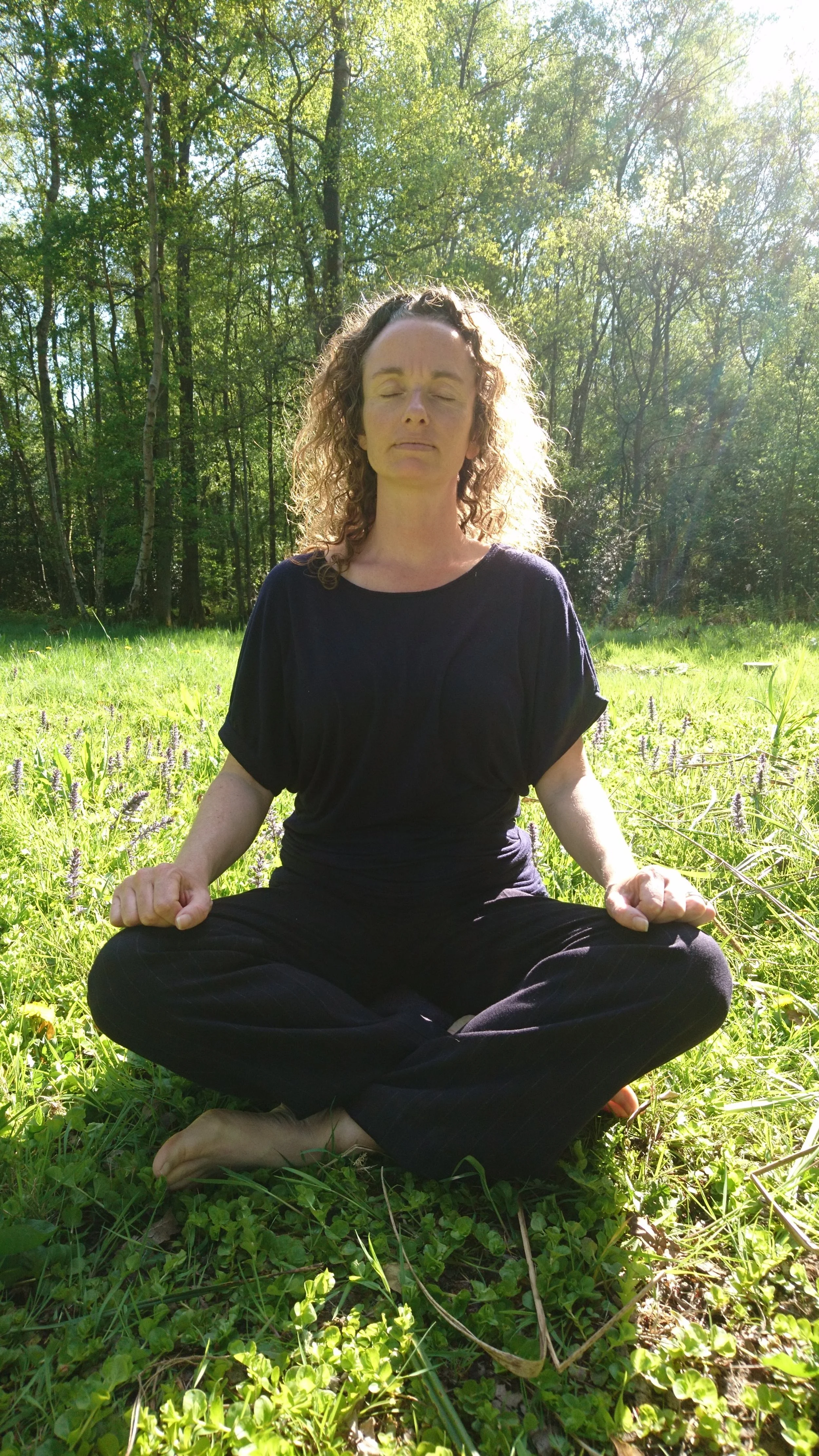 A woman with curly hair sitting cross-legged on grass in a forest, eyes closed, meditating in bright sunlight.
