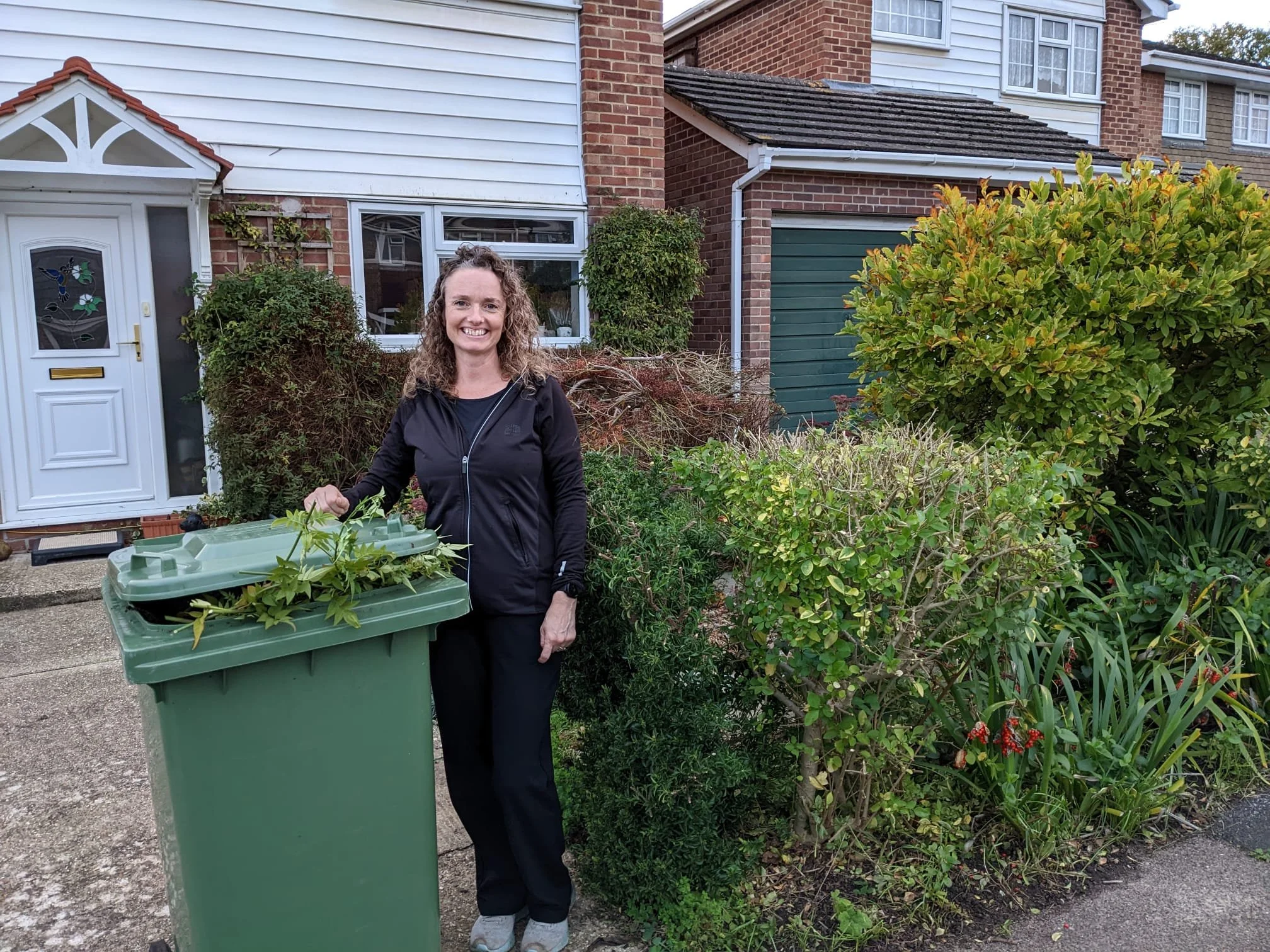A woman standing next to a green trash bin filled with garden clippings in front of a residential home with shrubs and plants.