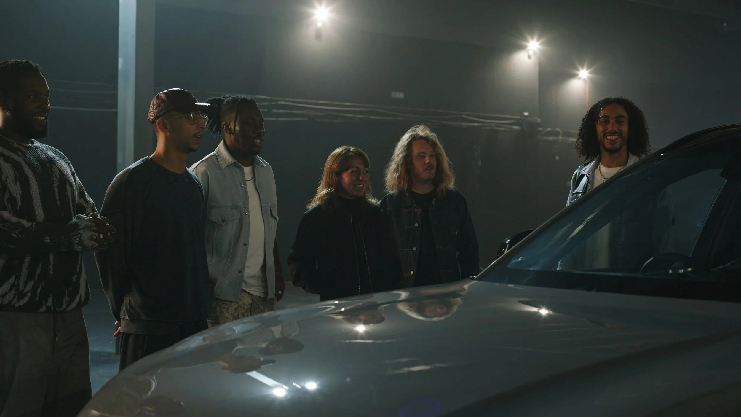 Group of six people standing outdoors at night, looking at a car, with streetlights overhead.