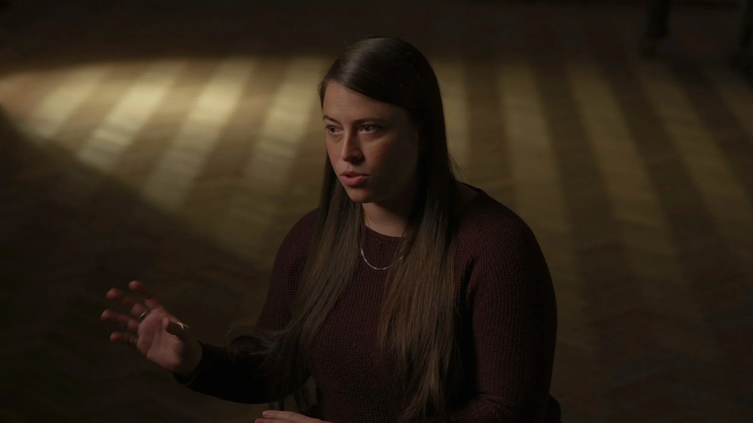 A woman with long brown hair, wearing a dark red sweater and a silver necklace, is speaking and gesturing with her right hand in a dimly lit room with a wooden floor and a shadow pattern.